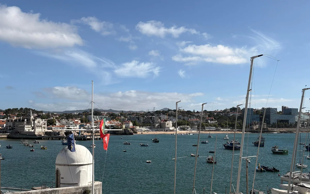 Mástiles de veleros y una pequeña torreta blanca con cúpula frente a una bahía de barcos amarrados y una localidad