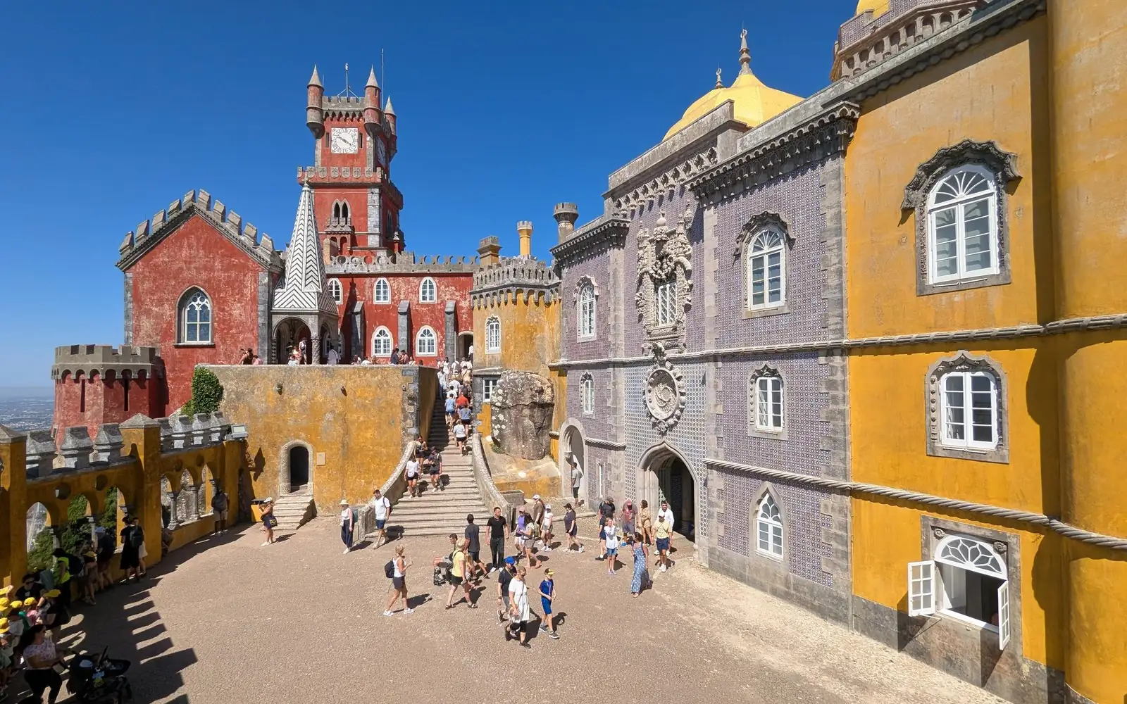 Besucher überqueren den Innenhof des Pena Palace in Sintra, unter einem roten Uhrturm und gelben Mauern