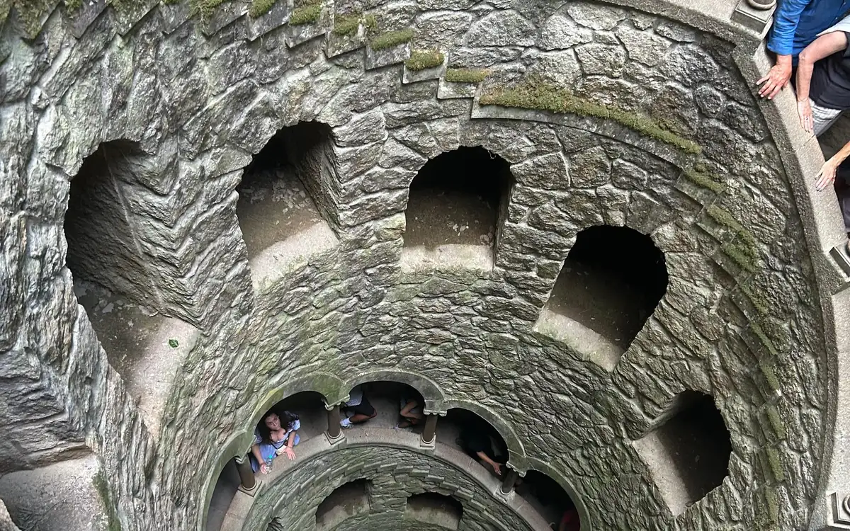 Vista hacia abajo de un pozo de piedra en espiral con aberturas en arco y visitantes en los niveles inferiores