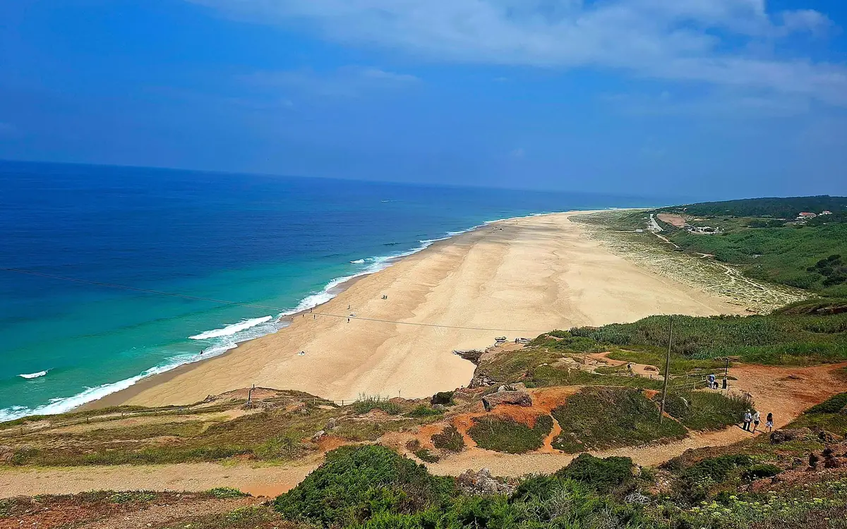 Una lunga spiaggia sabbiosa e mare turchese sotto scogliere verdi coperte di arbusti, con escursionisti su un sentiero