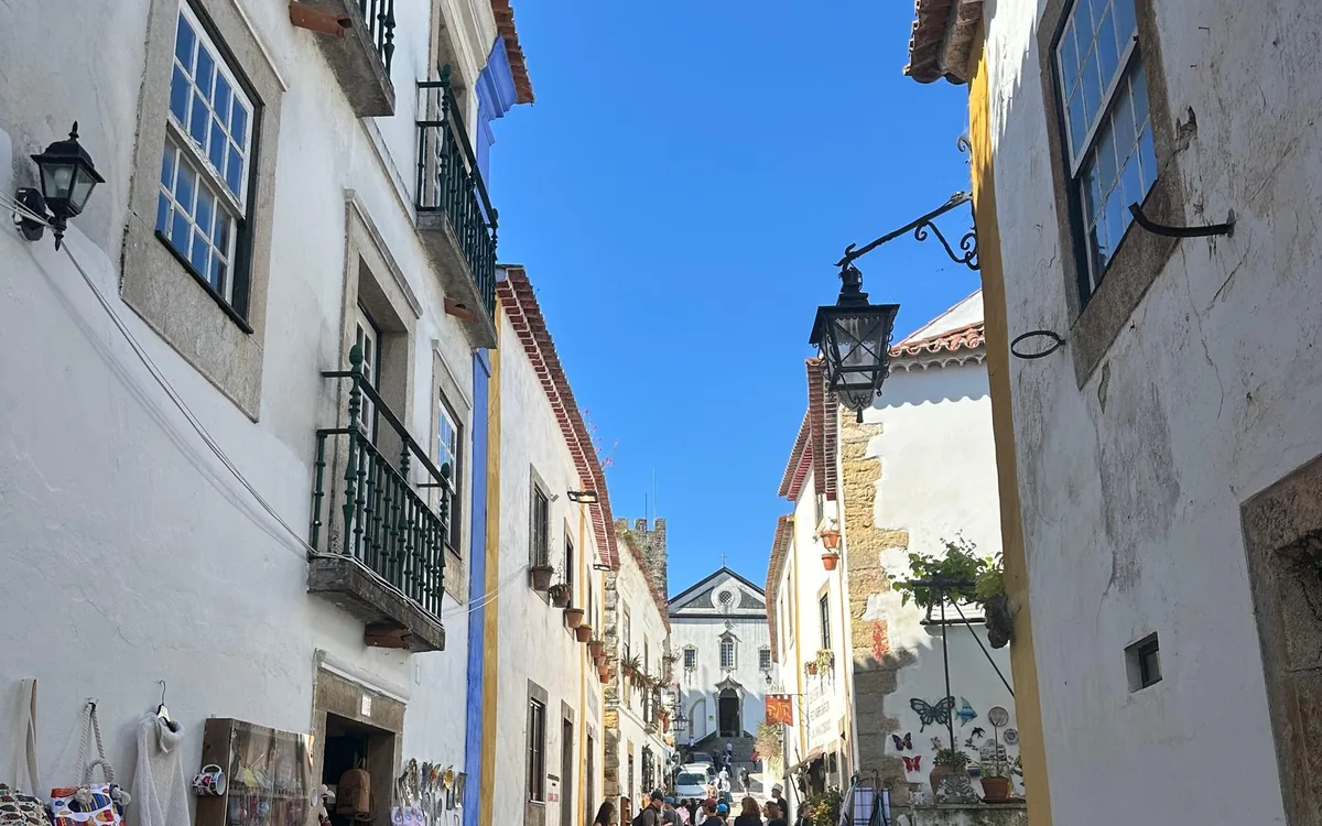 Uma rua estreita de casas brancas com lanternas de ferro, varandas e uma igreja ao fundo