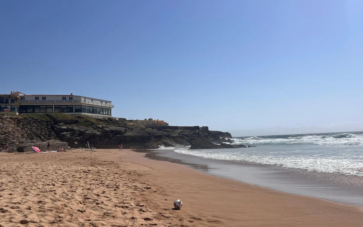 Una amplia playa de arena con espuma blanca a un lado y rocas oscuras con un edificio al otro
