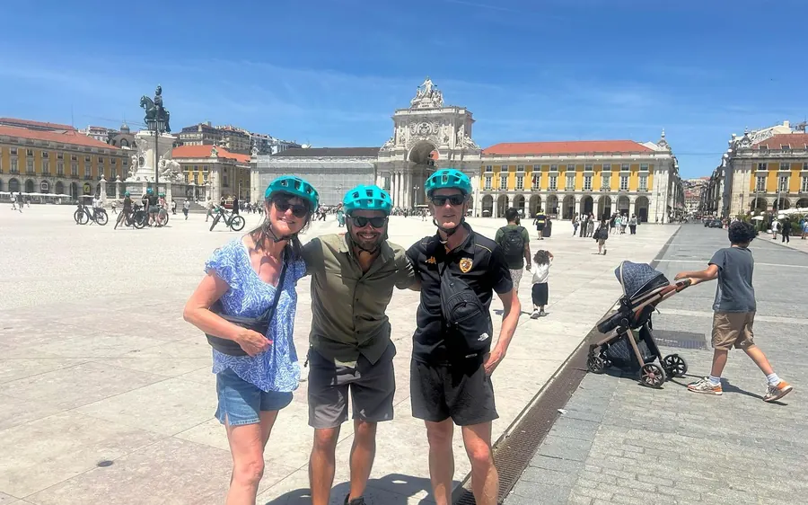Drei Fahrer mit Fahrradhelmen auf einem weiten Stadtplatz mit einem Bogen im Hintergrund